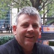 A head and shoulders photo of a man with short grey hair wearing a navy polo shirt with a tree in the background.