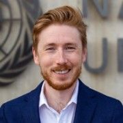 A head and shoulders of a man with ginger hair and a beard wearing a blue suit jacket and with a concrete wall and the edge of the UN logo in the background.