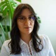 A head and shoulders photo of a woman with long dark hair, glasses, a white shirt and a plant in the background.